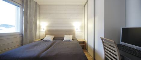 A bedroom with a bed, bedside tables, a desk with a computer, and a window.