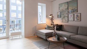 Een moderne woonkamer met een grijze bank, een ronde salontafel en een vloerlamp. Door de glazen deuren is een balkon zichtbaar met een tafel en stoelen.