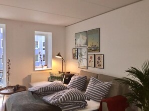 Een gezellige woonkamer met een bank, een bijzettafeltje, een lamp en een raam met uitzicht op gebouwen.