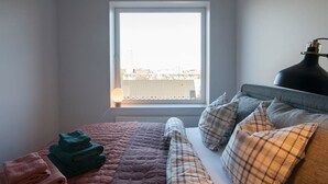 Een slaapkamer met een bed, kussens, een lamp en een raam met uitzicht op gebouwen.