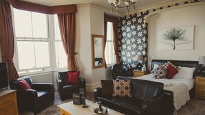 A hotel room with a bed, two black leather sofas, a wooden coffee table, a television, and a large window with curtains. - The Crown Hotel (Morecambe)