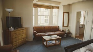 A room with a brown leather sofa, a wooden coffee table with magazines, a TV on a wooden cabinet, and a bathroom visible through an open door. - The Crown Hotel (Morecambe)