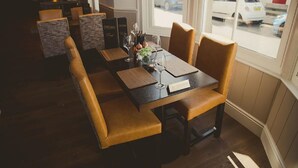 A restaurant with a long table, cushioned chairs, and a view of the street through large windows. - The Crown Hotel (Morecambe)