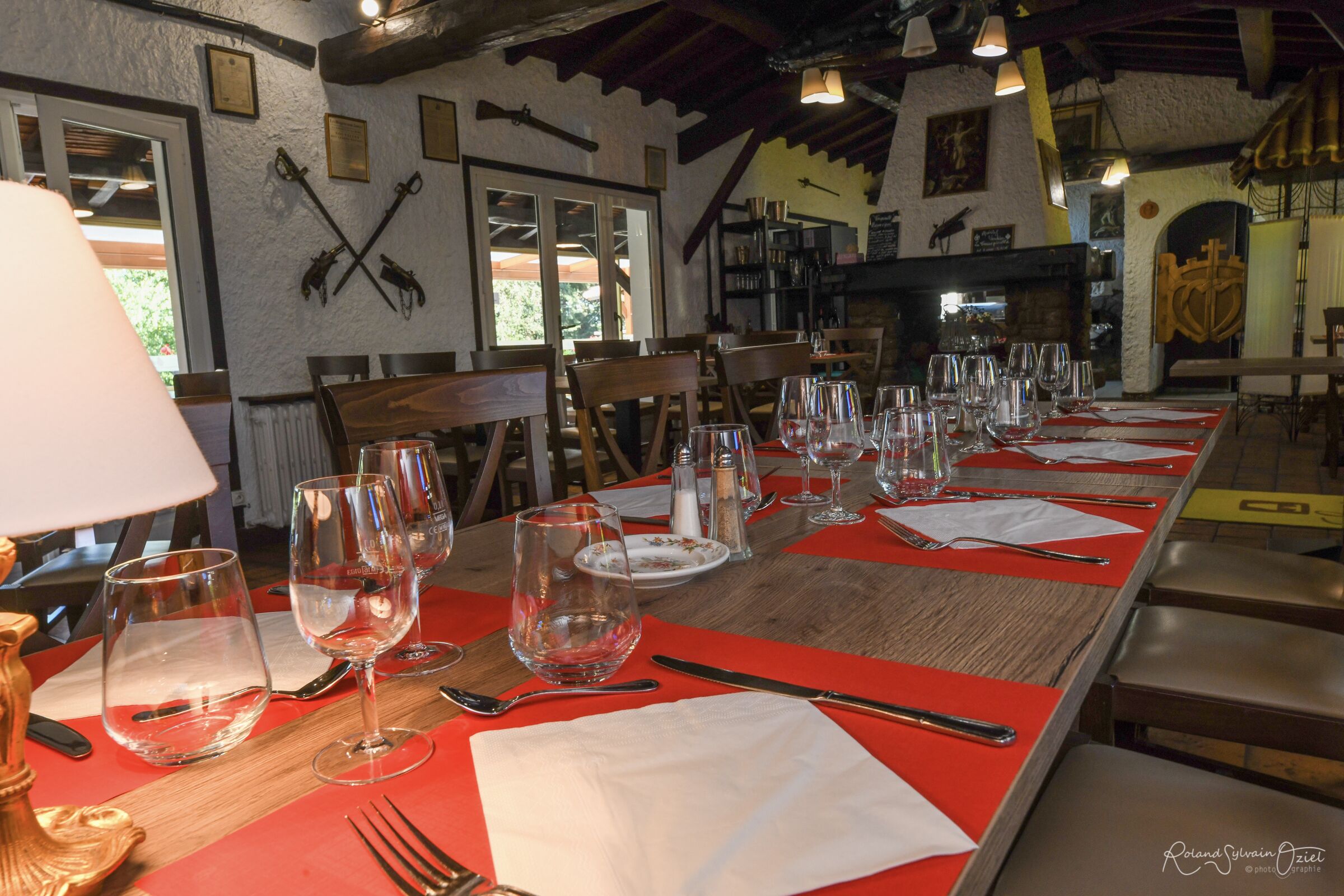 A dining table set with wine glasses, plates, and cutlery, surrounded by wooden chairs in a rustic interior with exposed beams and a fireplace.