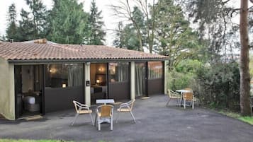 Um pequeno edifício térreo, com telhado em telhas e janelas amplas. Possui um espaço de pátio com área de estar externa e uma mesa.