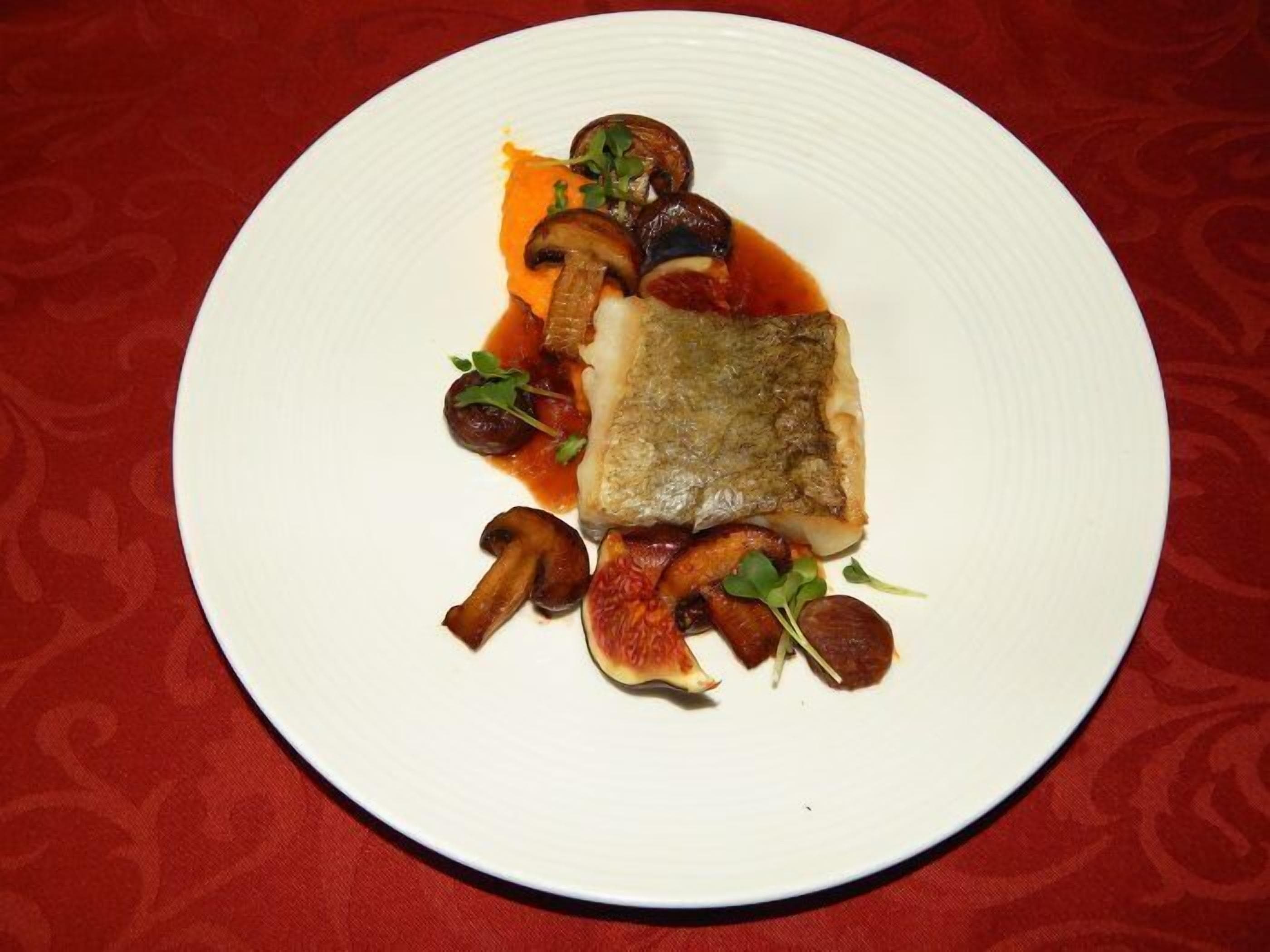 A white plate with a piece of fish, mushrooms, and a sauce on a red tablecloth.
