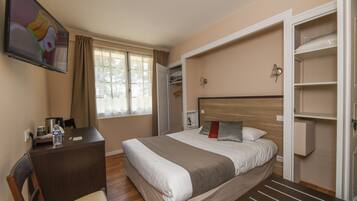 Quarto de hotel com cama, escrivaninha, cadeira, televisão, guarda-roupa e janela com cortinas.