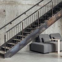 A staircase with metal railings and a concrete wall. There is a gray sofa with a small table beside it.