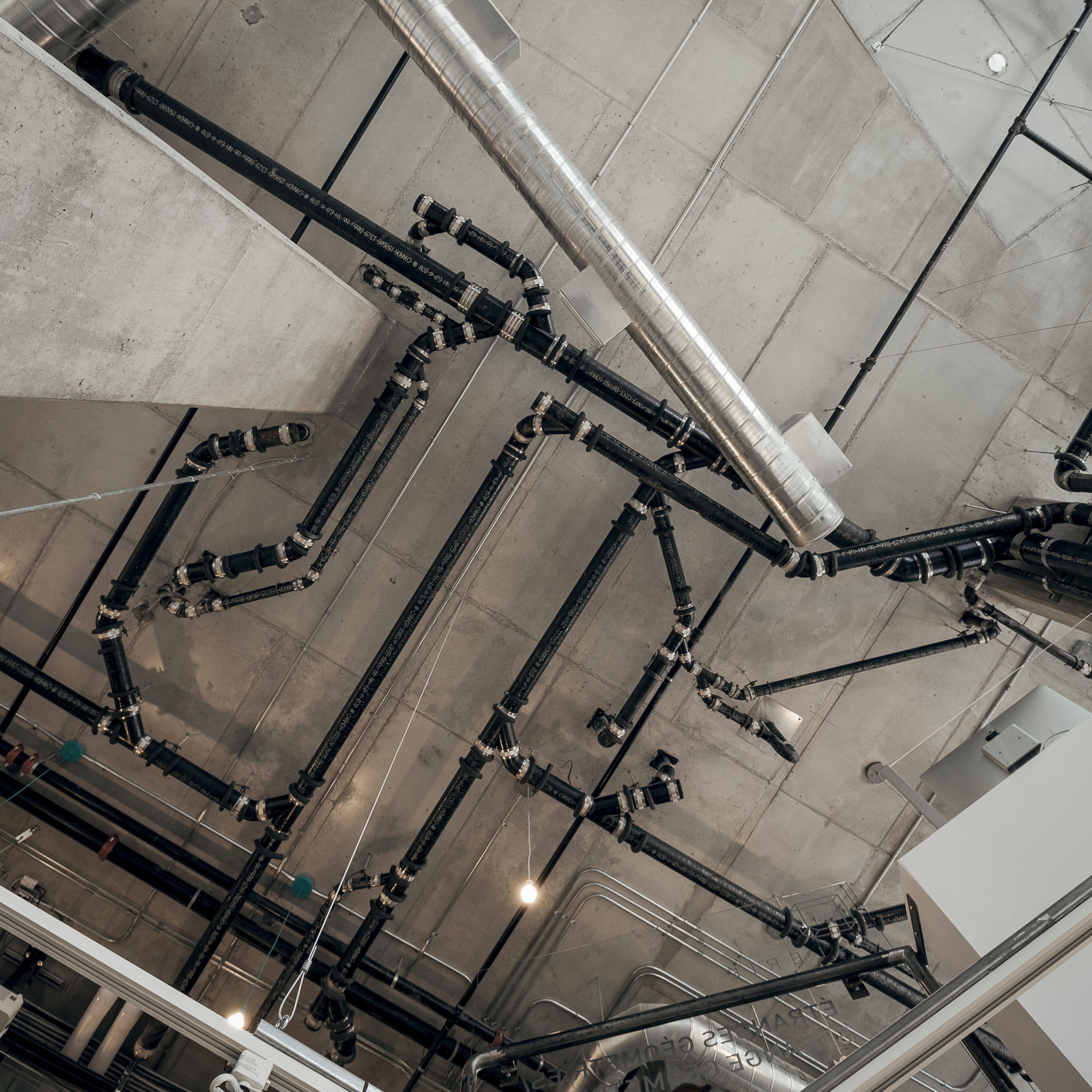 A complex network of black pipes and valves on a concrete ceiling, likely part of a building's infrastructure.
