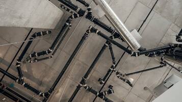 A complex network of black pipes and valves on a concrete ceiling, likely part of a building's infrastructure.