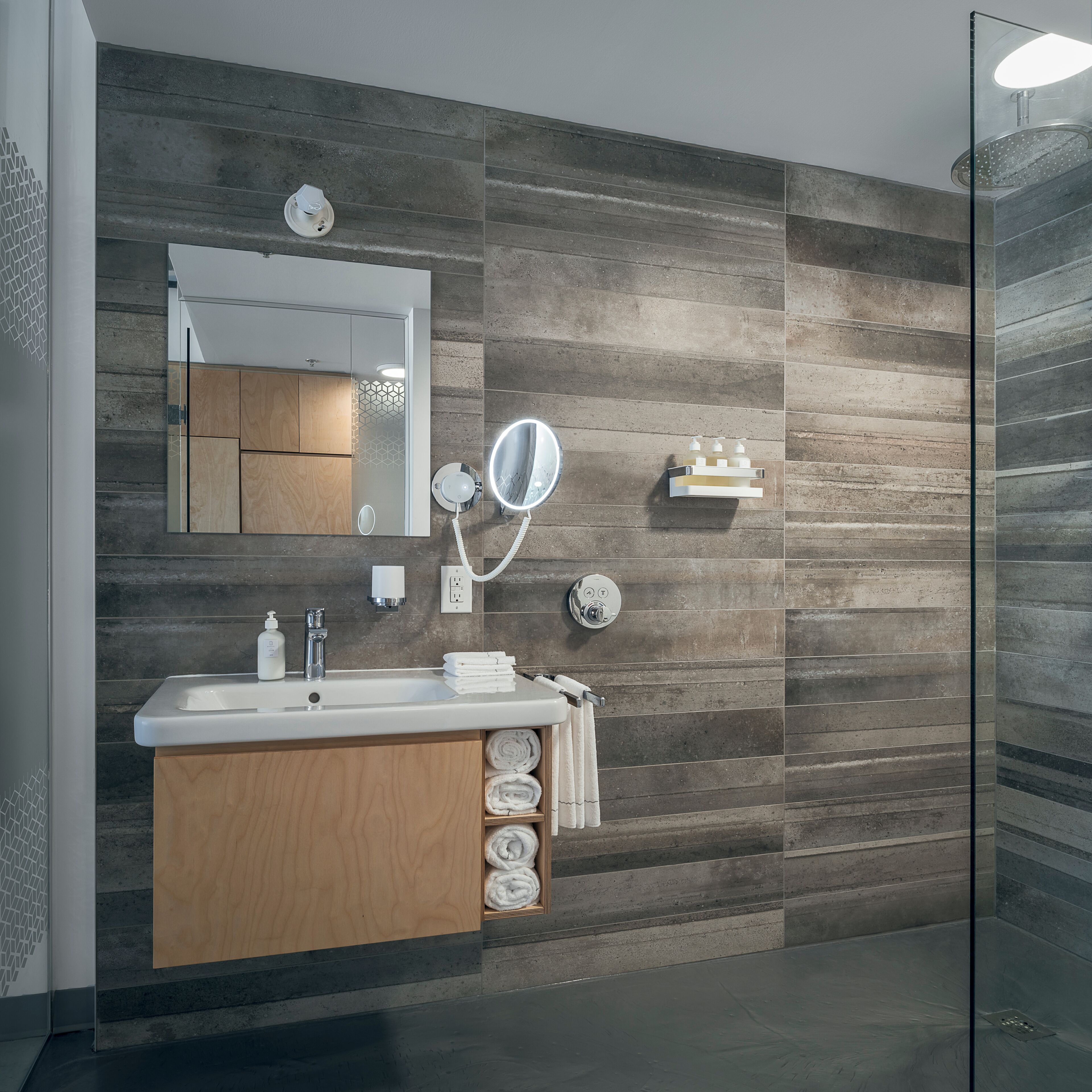A modern bathroom with a wooden vanity, a large mirror, a built-in shelf with toiletries, and a glass shower enclosure.