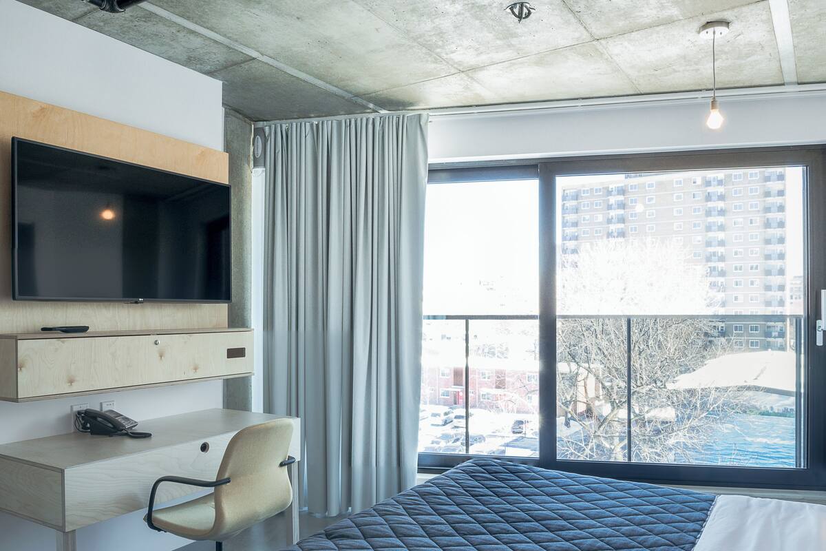 A modern hotel room with a large bed, a desk with a chair, a TV mounted on the wall, and a window with a view of buildings and trees.