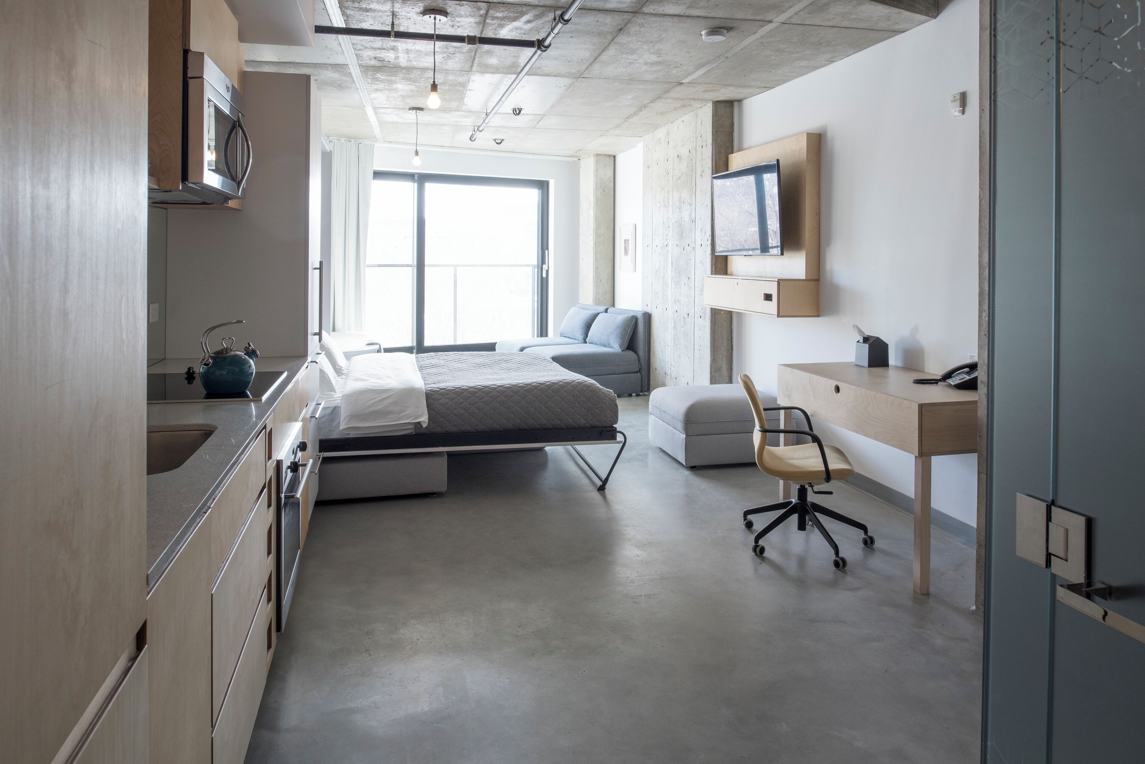 Une chambre d’hôtel moderne équipée d’une kitchenette, d’un lit, d’un bureau et d’une grande fenêtre.