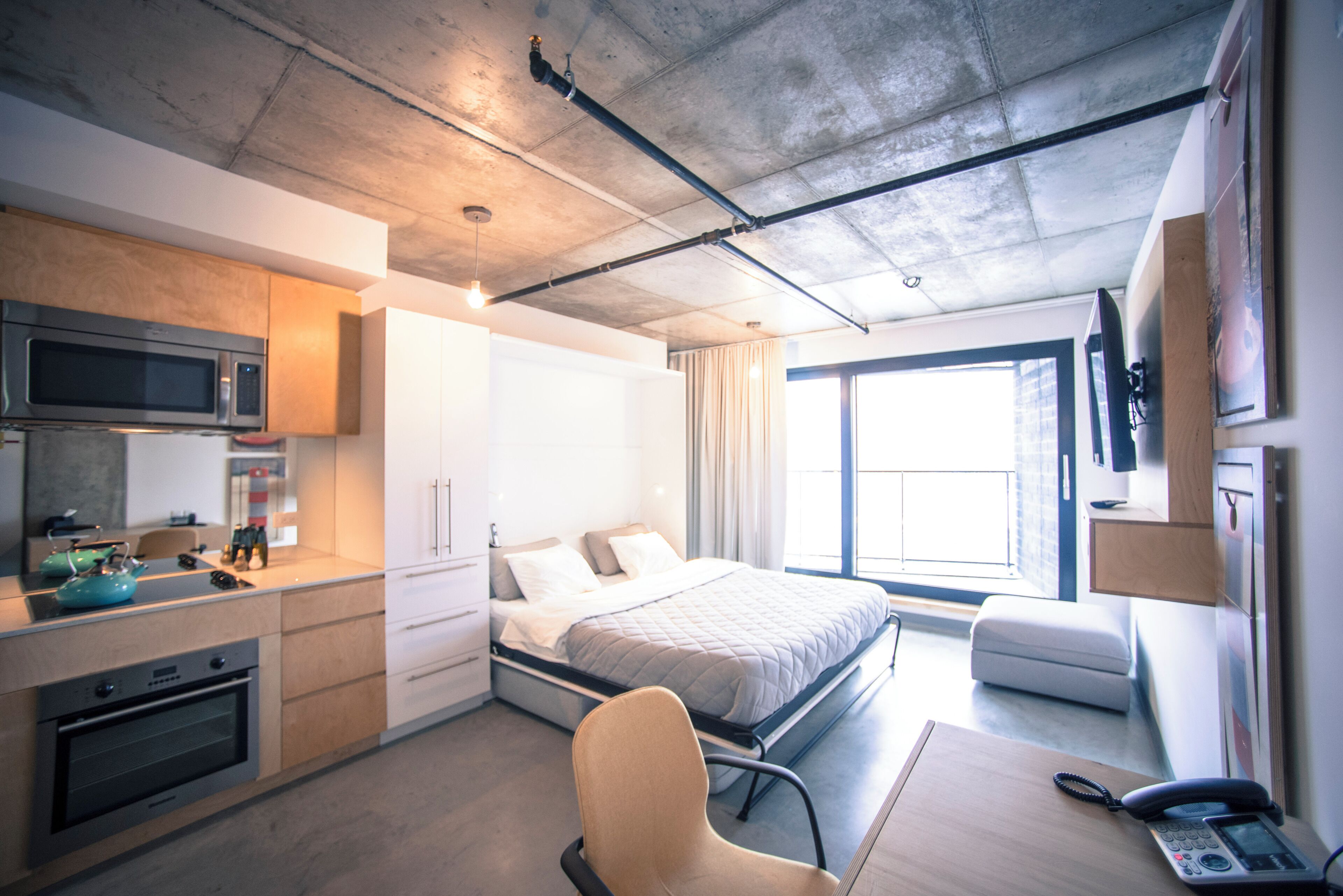 A modern hotel room with a kitchenette, a bed, a chair, and a desk with a phone.