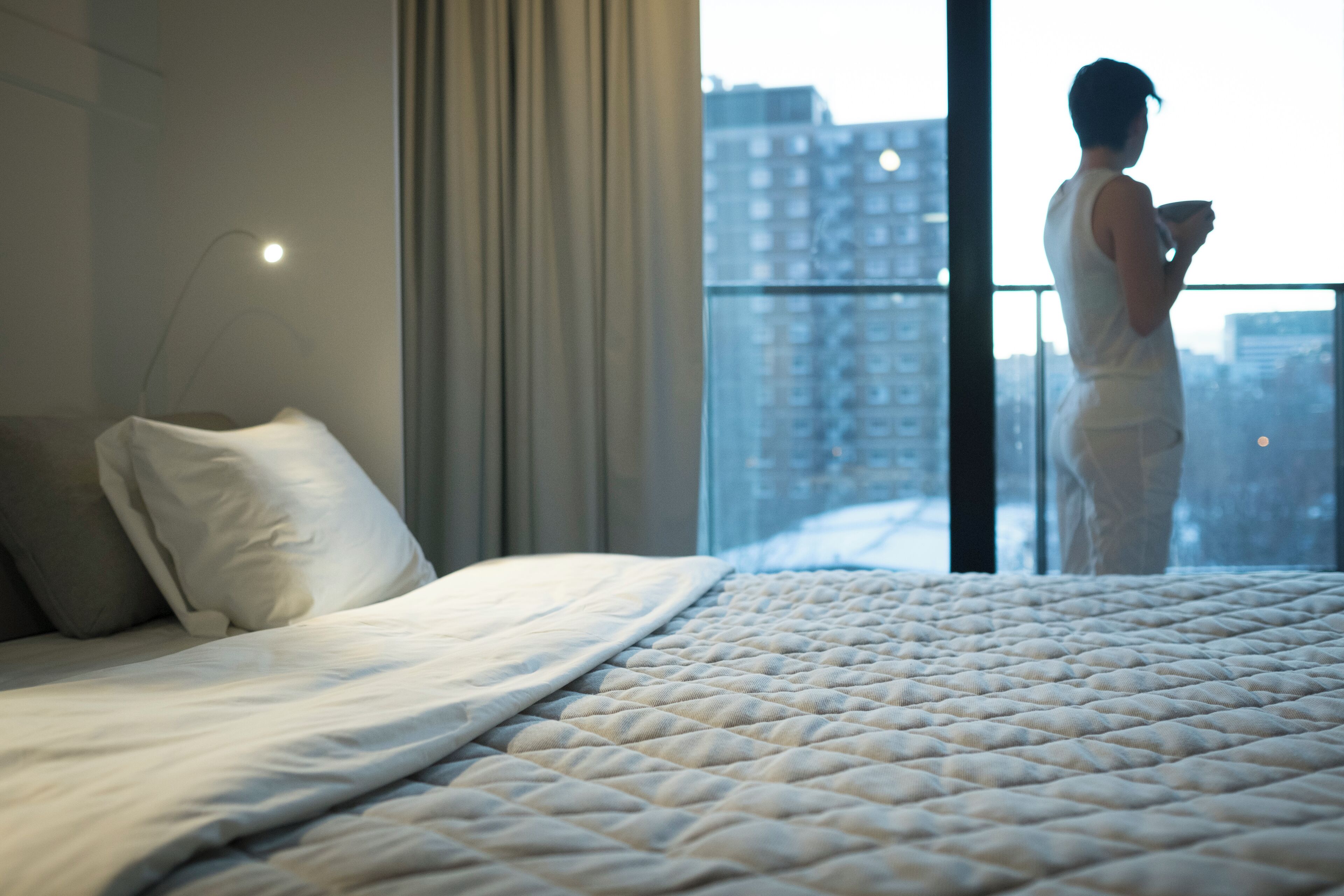 A hotel room with a large bed, a window with a view of a cityscape, and a person standing by the window.