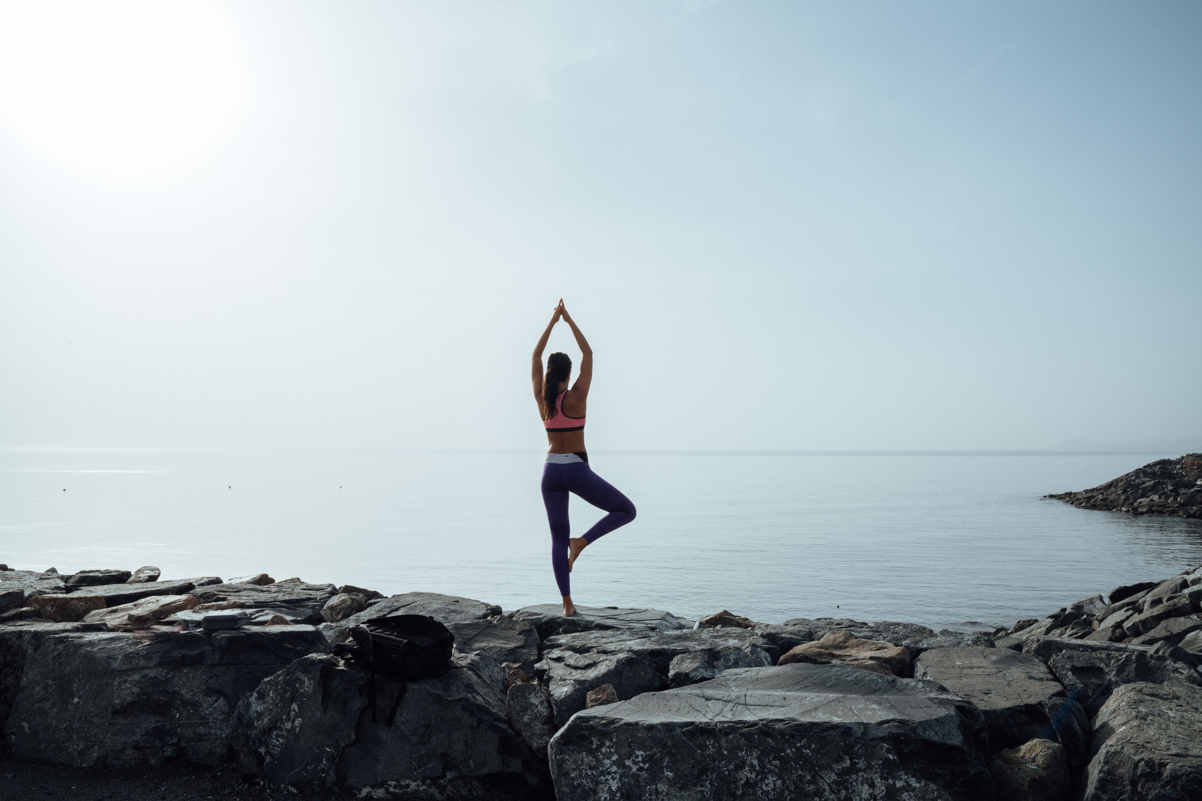 Una persona realizando una postura de yoga en una playa rocosa con agua tranquila al fondo.