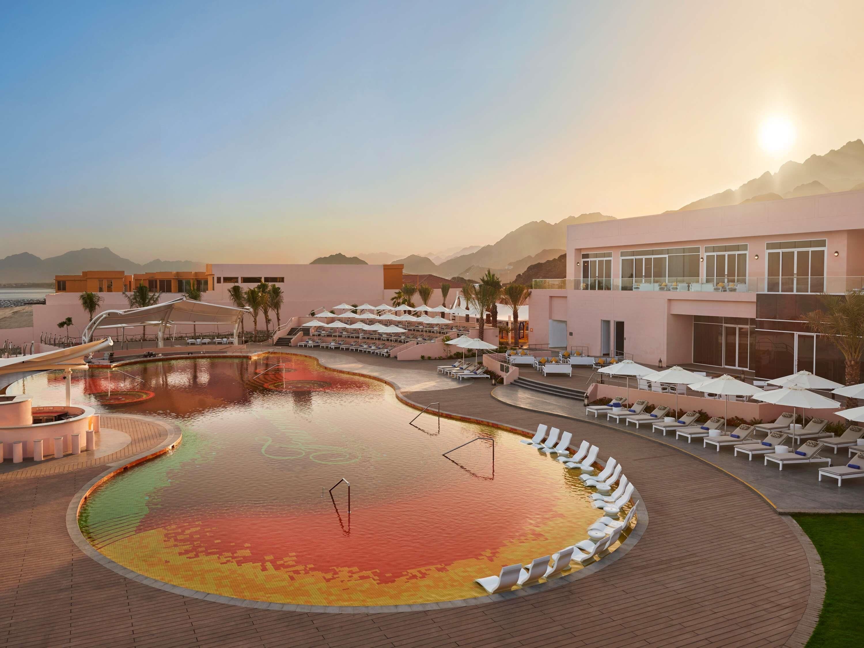 Une piscine d’hôtel avec des chaises longues et des parasols, entourée d’immeubles et de montagnes.