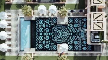 An aerial view of a rooftop pool area with a geometric tile pattern, lounge chairs, umbrellas, and potted plants.