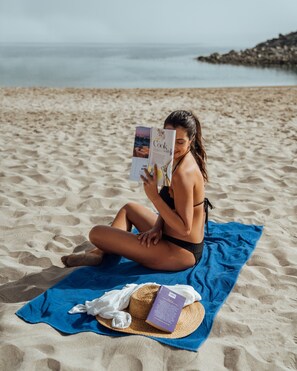 Une personne assise sur une serviette de plage, en train de lire un livre.