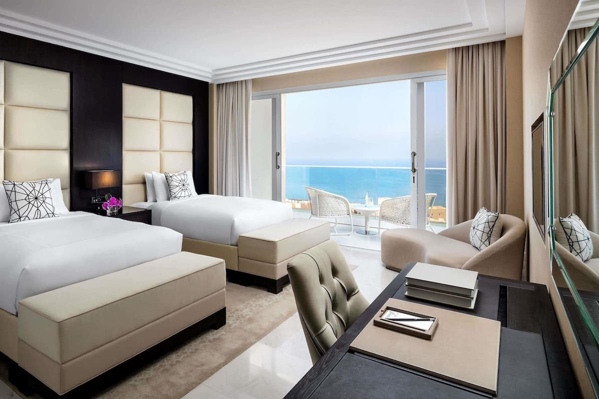 Une chambre d’hôtel avec deux lits, un balcon donnant sur la mer et un coin repas avec une table et des chaises.