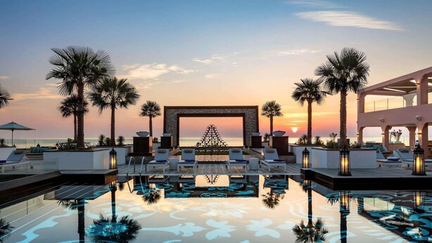 Zona de piscina con sillones reclinables, palmeras y vistas al atardecer.