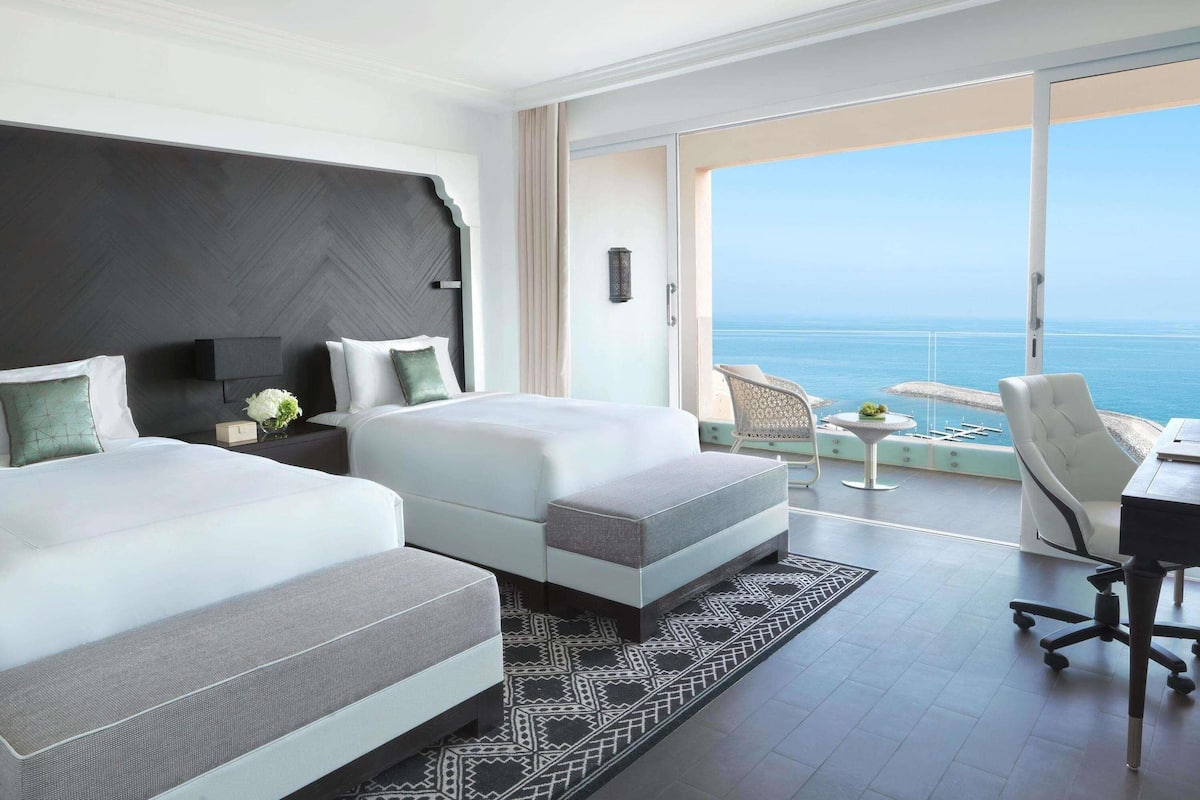 Une chambre d’hôtel moderne avec deux lits, un balcon donnant sur la mer et un bureau avec une chaise.