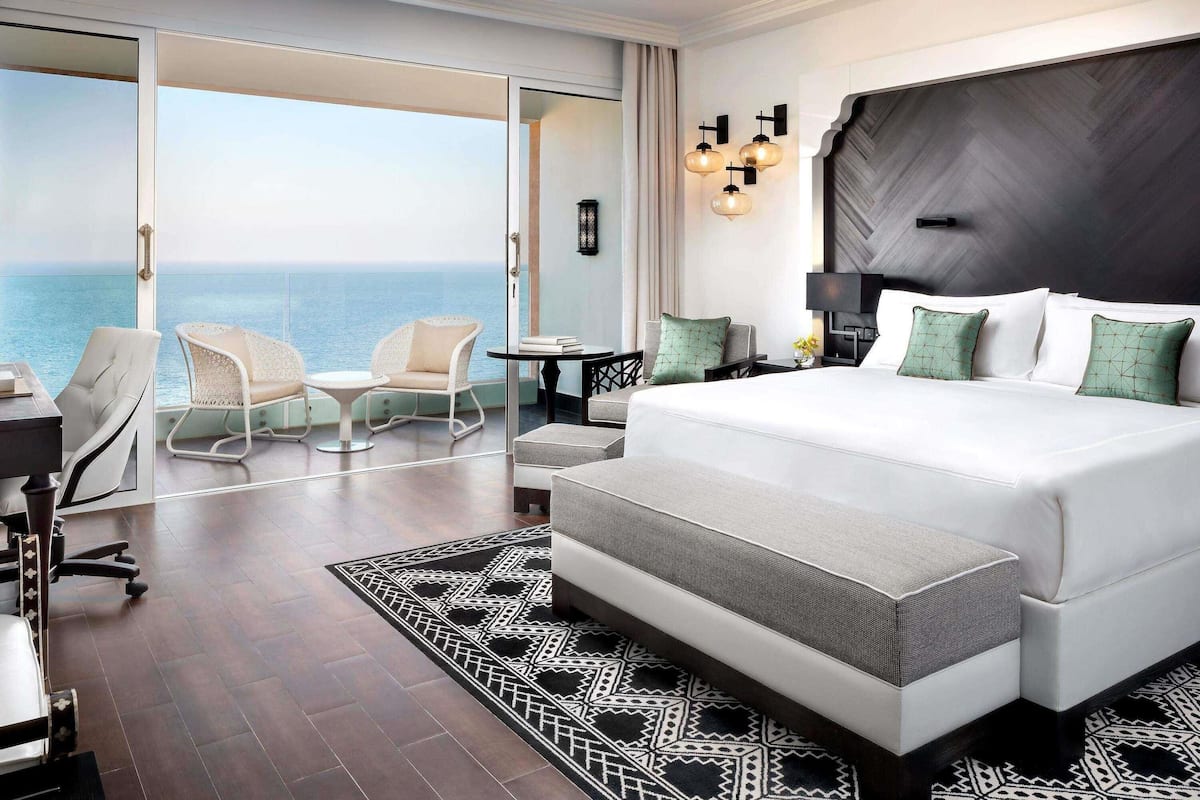 Une chambre d’hôtel avec un grand lit, un balcon donnant sur la mer, un bureau avec une chaise et une petite table avec une lampe.