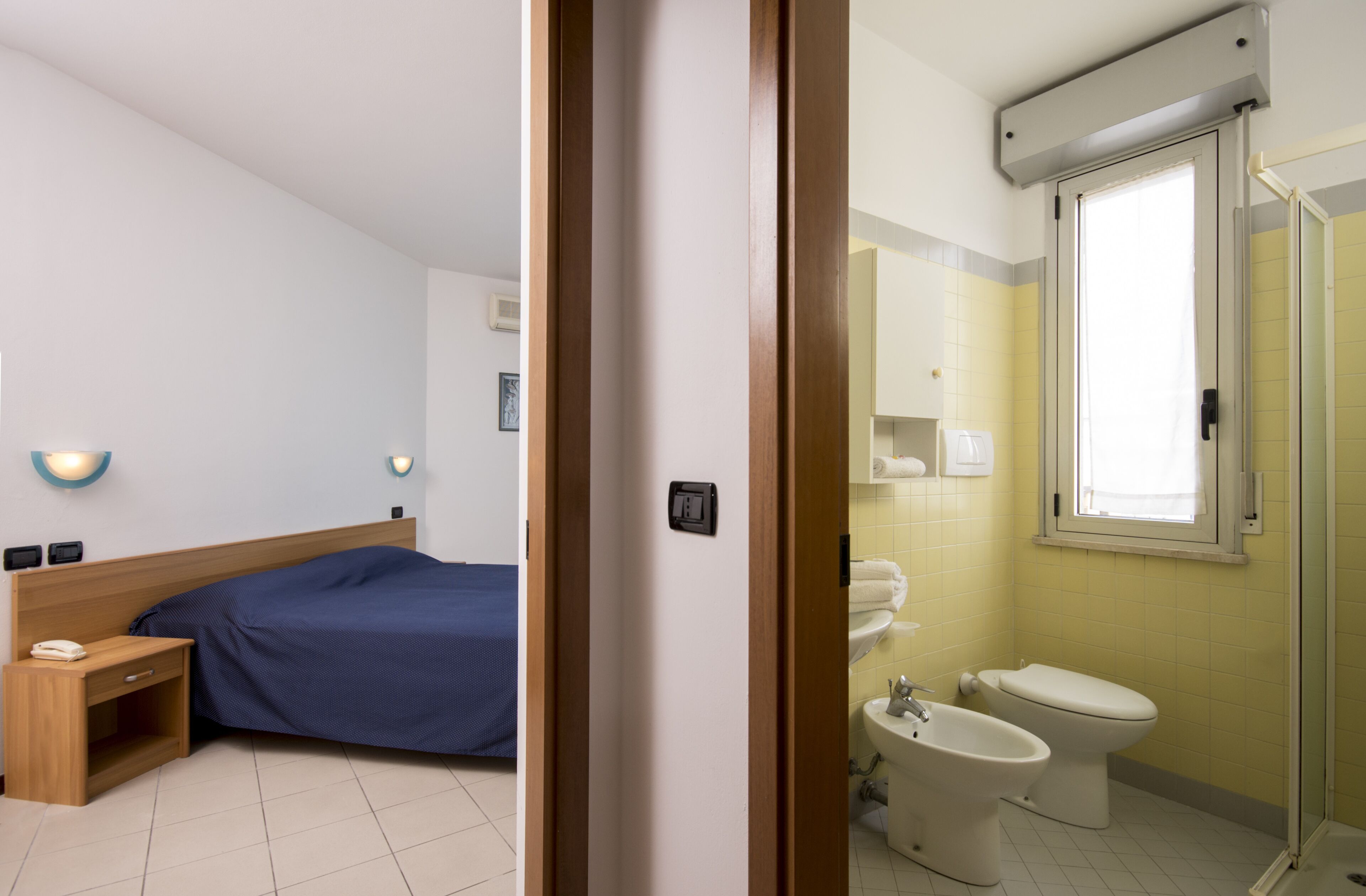 A bedroom with a bed, bedside table, and a bathroom with a toilet, bidet, and shower.