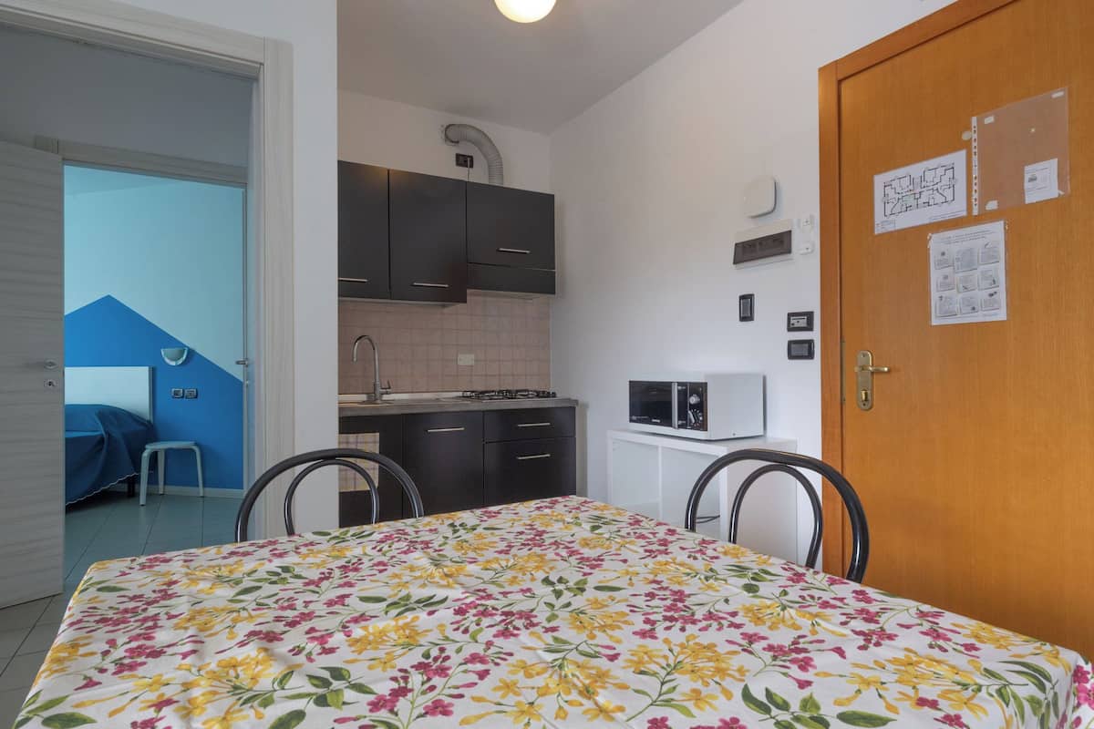 Ein kleines Zimmer mit einem Bett, einem Esstisch mit Stühlen, einer Küchenzeile und einer Tür mit Aushängen.