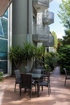 An outdoor patio with wicker furniture, a glass table, and a view of a building with a balcony. - Residence Villa Azzurra (Rivazzurra)