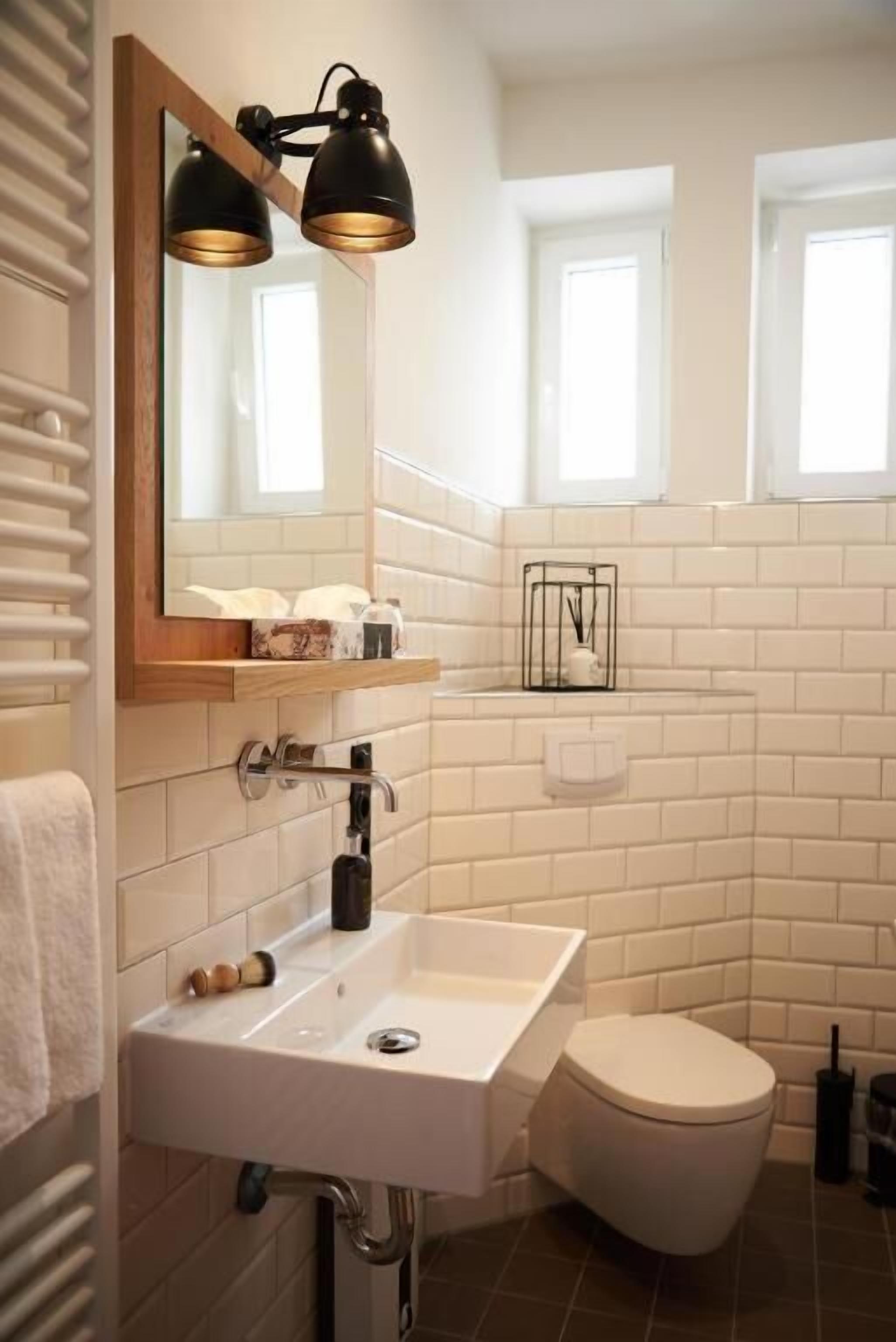 Un baño moderno con azulejos blancos tipo metro, un lavamanos blanco, un inodoro y un espejo con dos lámparas colgantes negras.