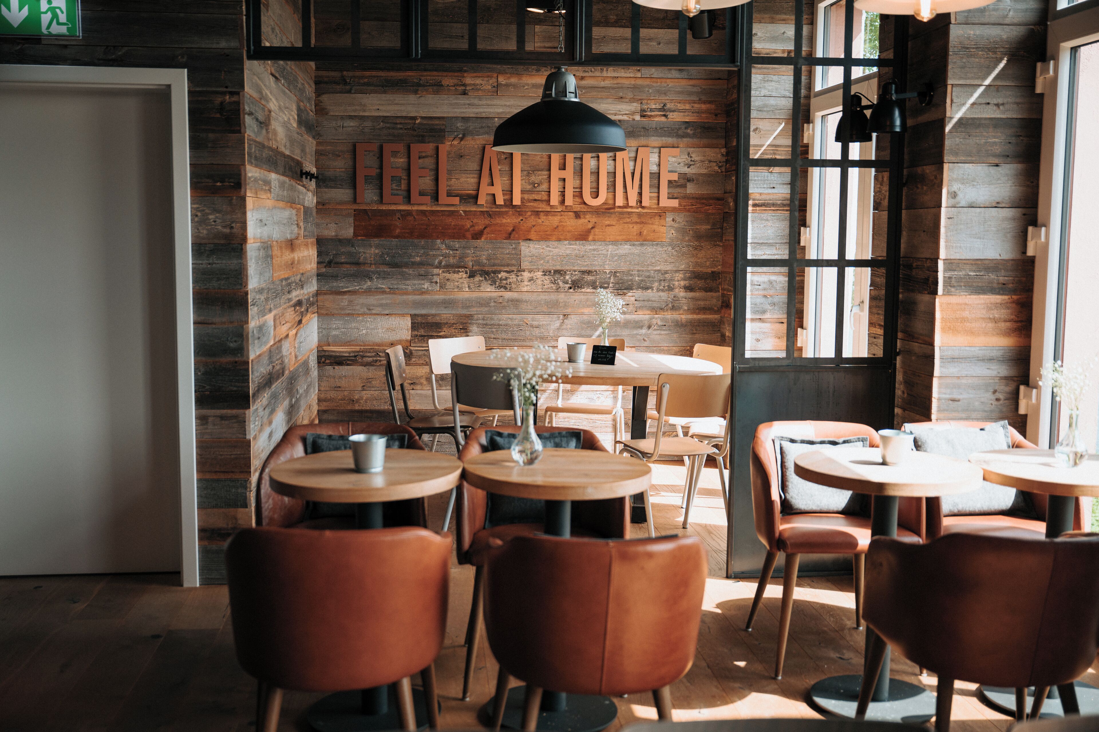 A cozy cafe with wooden walls, a 'FEEL AT HOME' sign, and round tables with leather chairs.