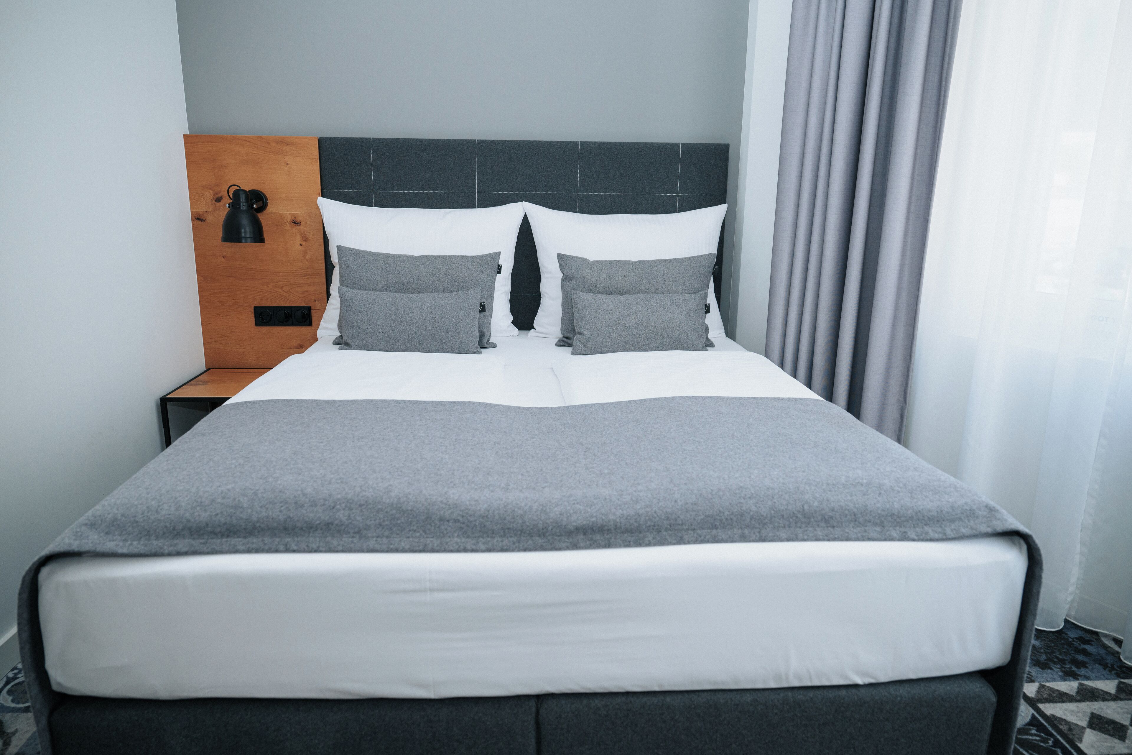 Una cama bien hecha con ropa de cama gris y blanca, un cabecero de madera y una mesita de noche con una lámpara.
