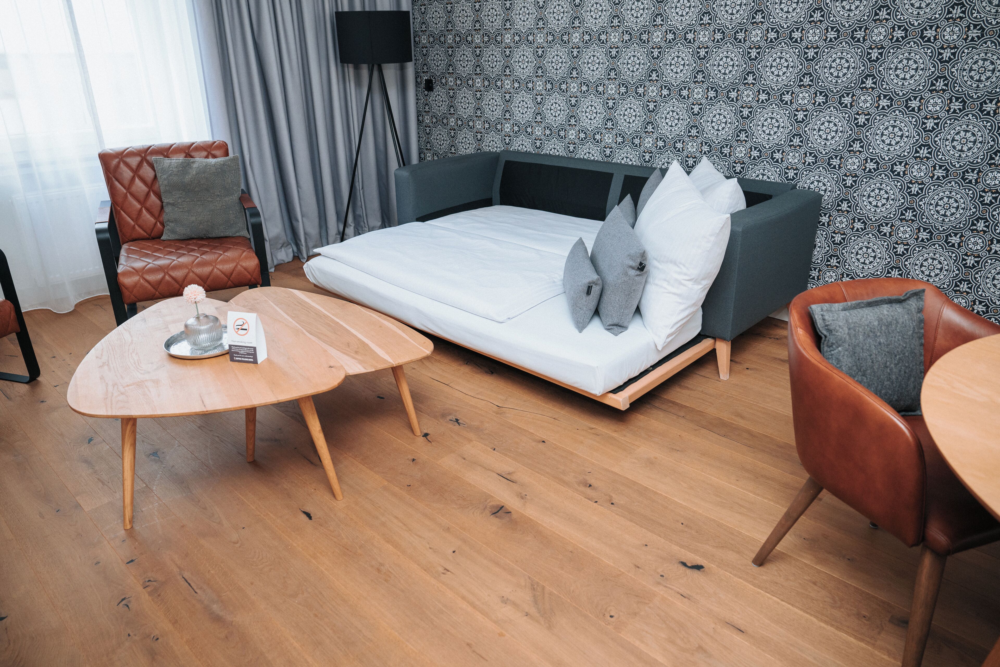 A modern room with a sofa bed, a wooden coffee table, a leather armchair, and a patterned wallpaper.