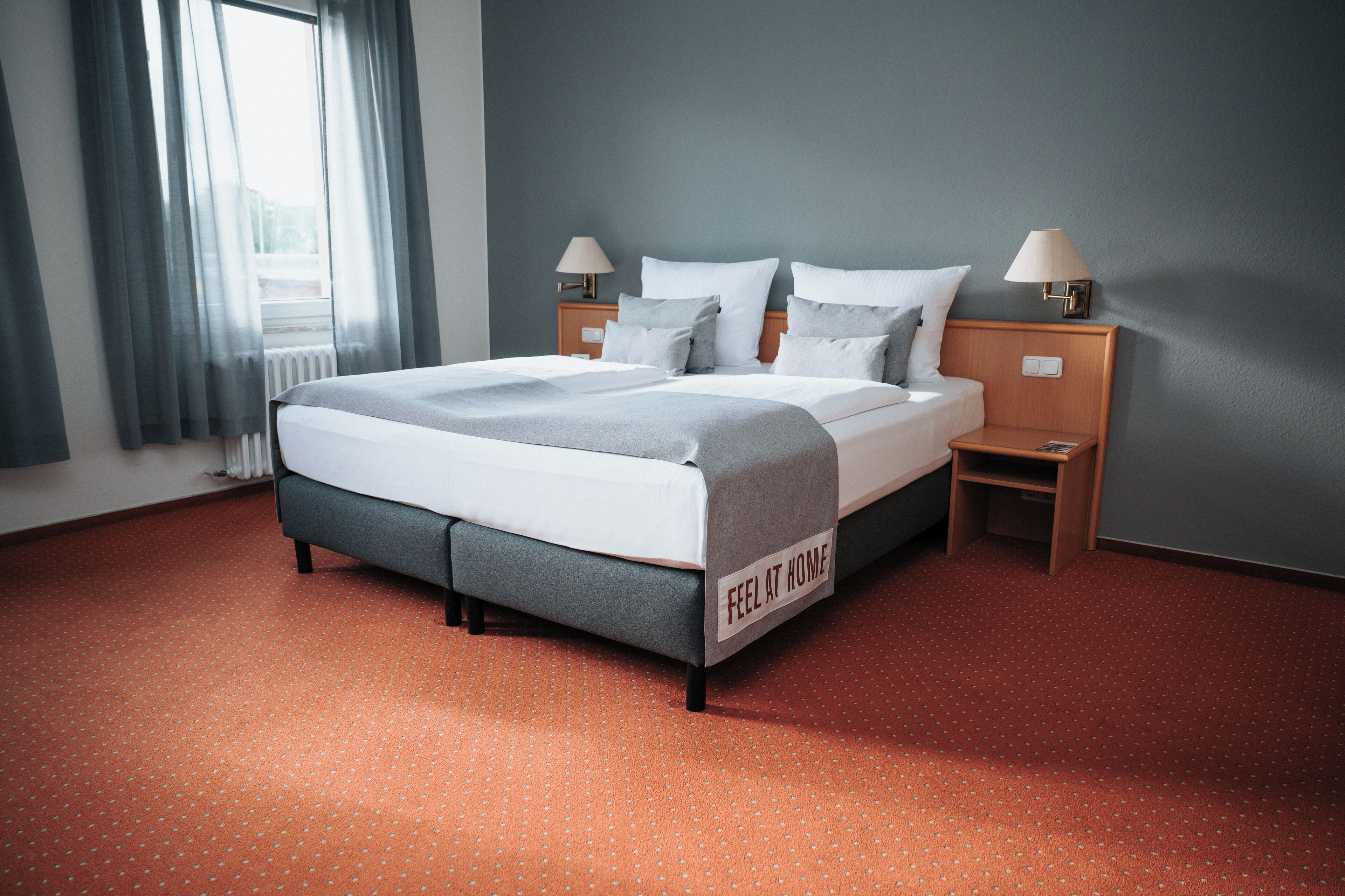 A neatly made bed with a grey and white duvet, two bedside tables with lamps, and a sign on the bed that reads "FEEL AT HOME".