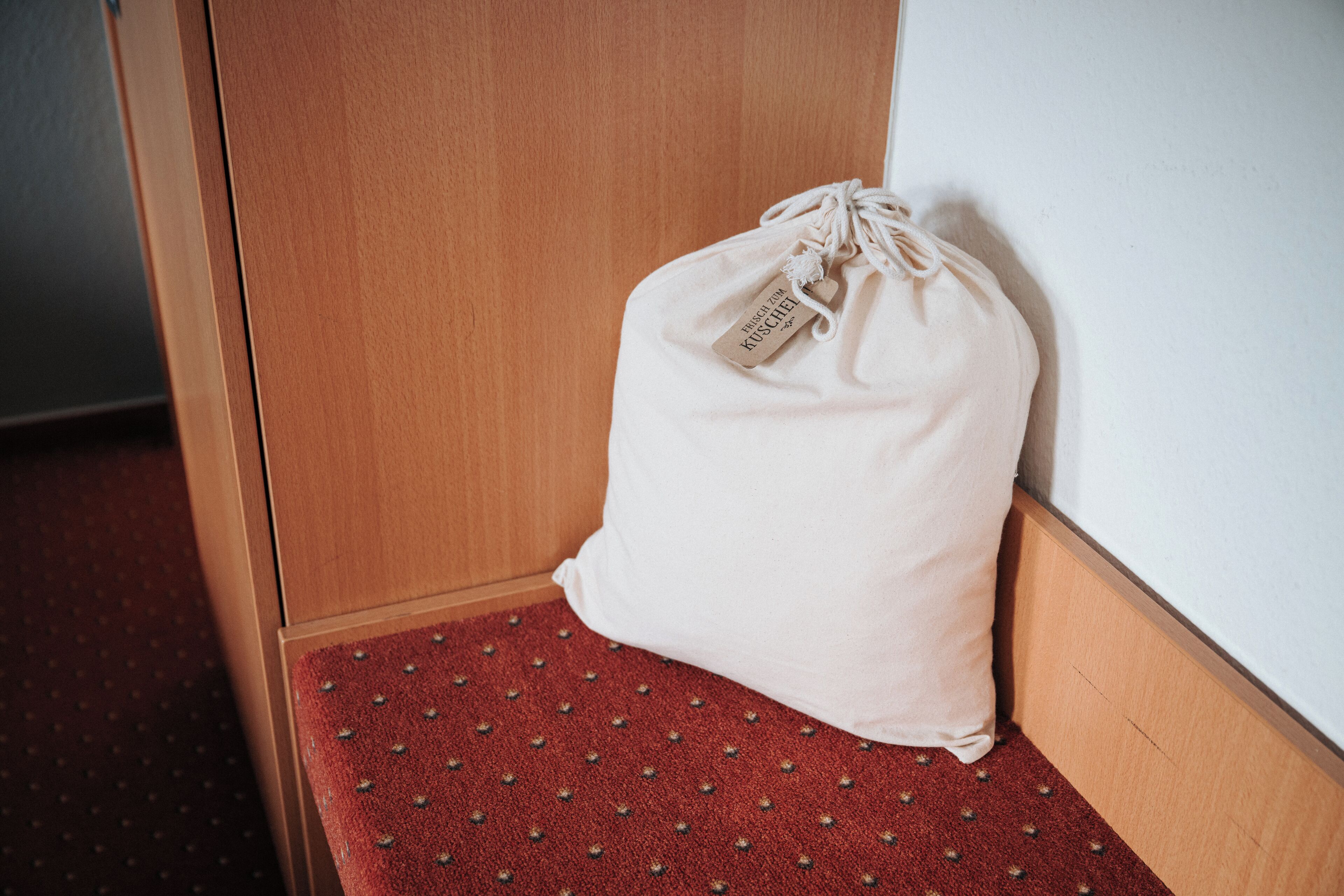 Una pequeña bolsa beige con cordón y una etiqueta sobre un banco con alfombra roja en un rincón de la habitación.