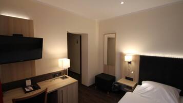 A hotel room with a bed, a desk with a chair, a television, a mirror, and a nightstand with a lamp.
