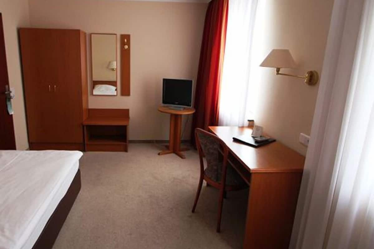 Habitación de hotel con cama, escritorio con silla, televisor, espejo y ventana con cortinas.