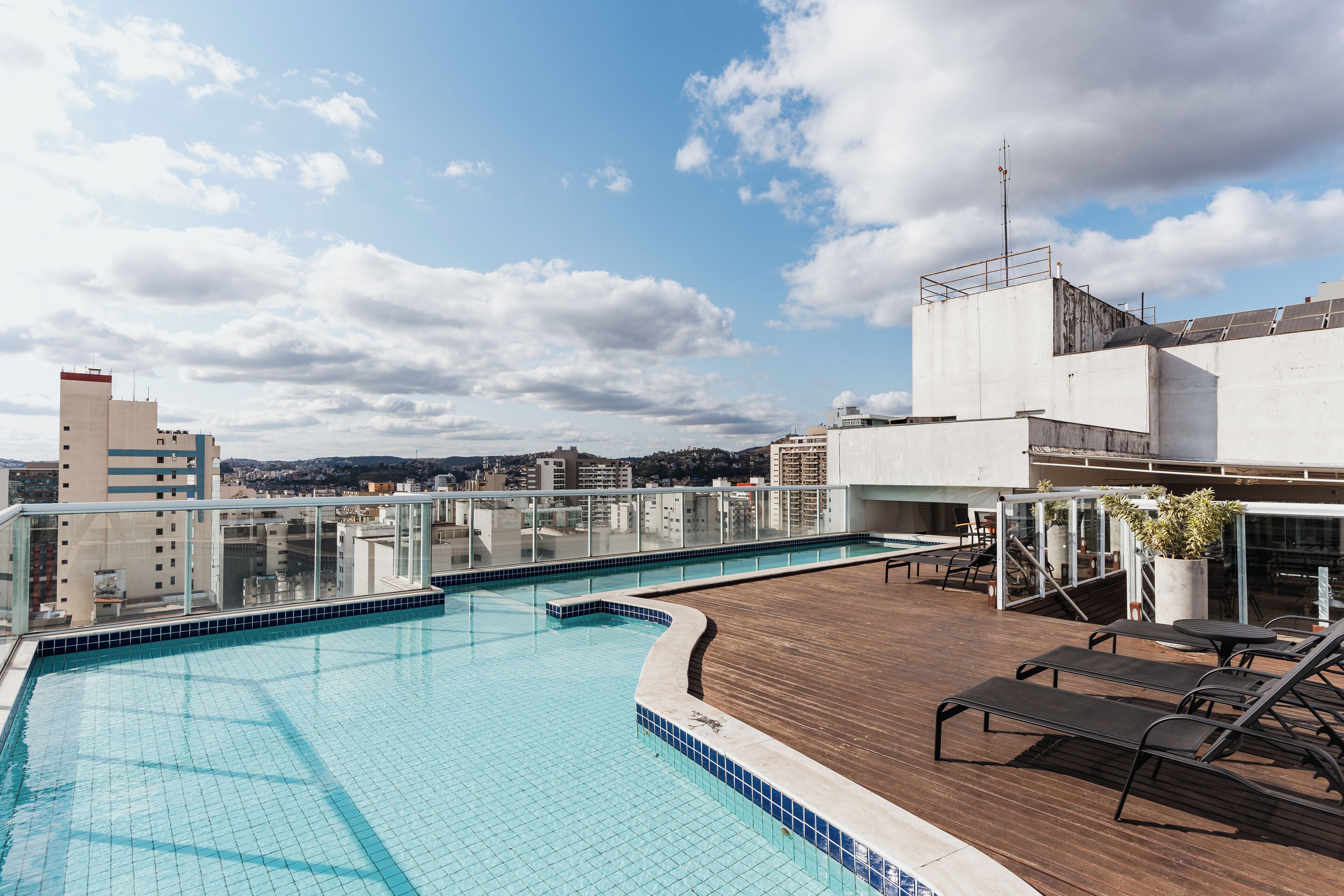 A rooftop swimming pool with a wooden deck and lounge chairs, surrounded by buildings and a clear sky.