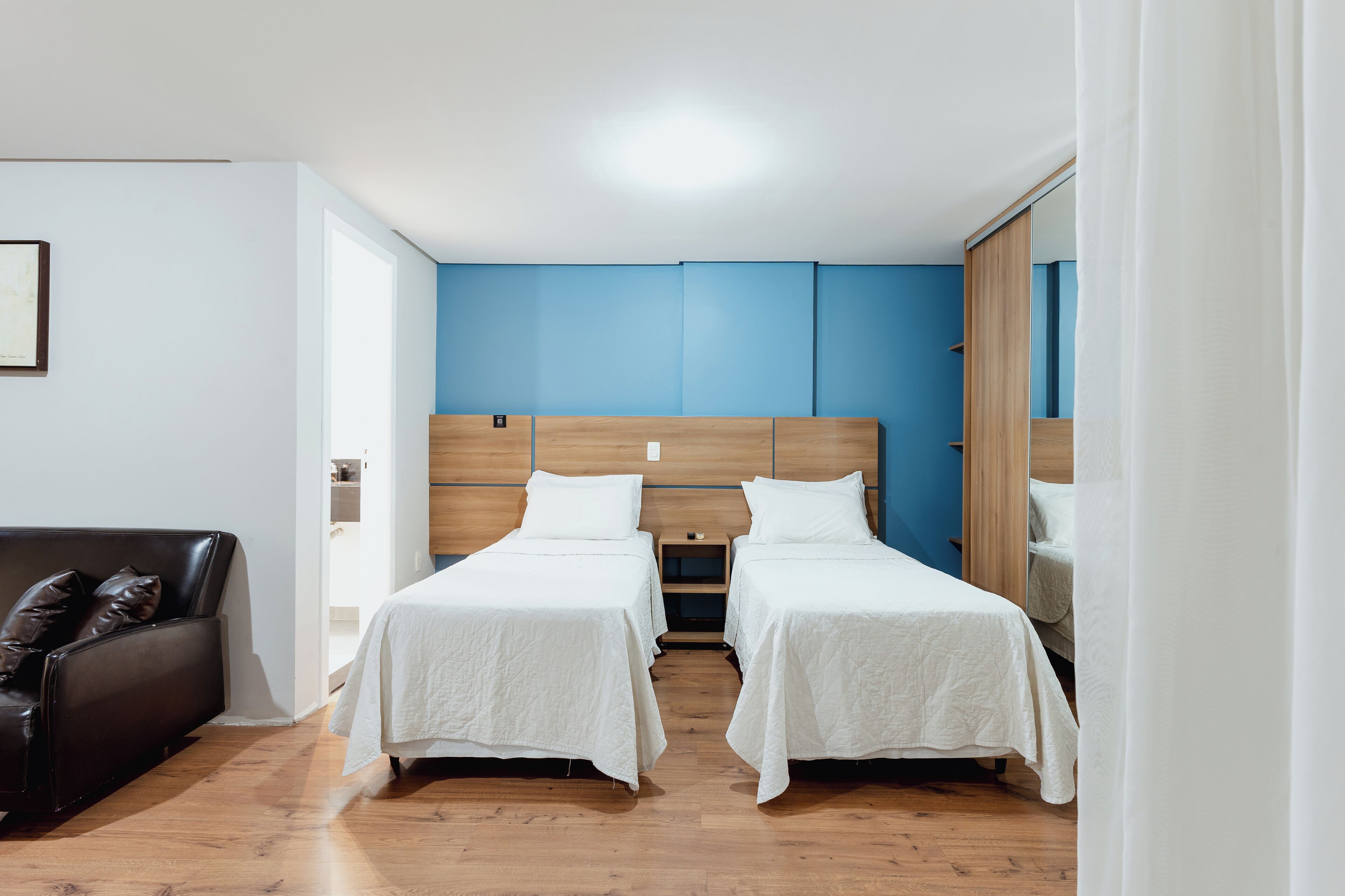 A hotel room with two single beds, a wooden headboard, a black leather sofa, and a large mirror.