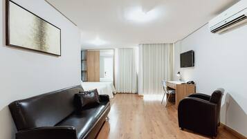 Moderni hotellihuone, jossa on musta nahkasohva, puinen työpöytä, litteänäyttöinen televisio ja suuri ikkuna, jossa on verhot.