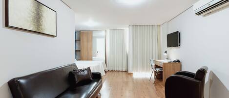 A modern hotel room with a black leather sofa, a wooden desk, a flat-screen TV, and a large window with curtains.