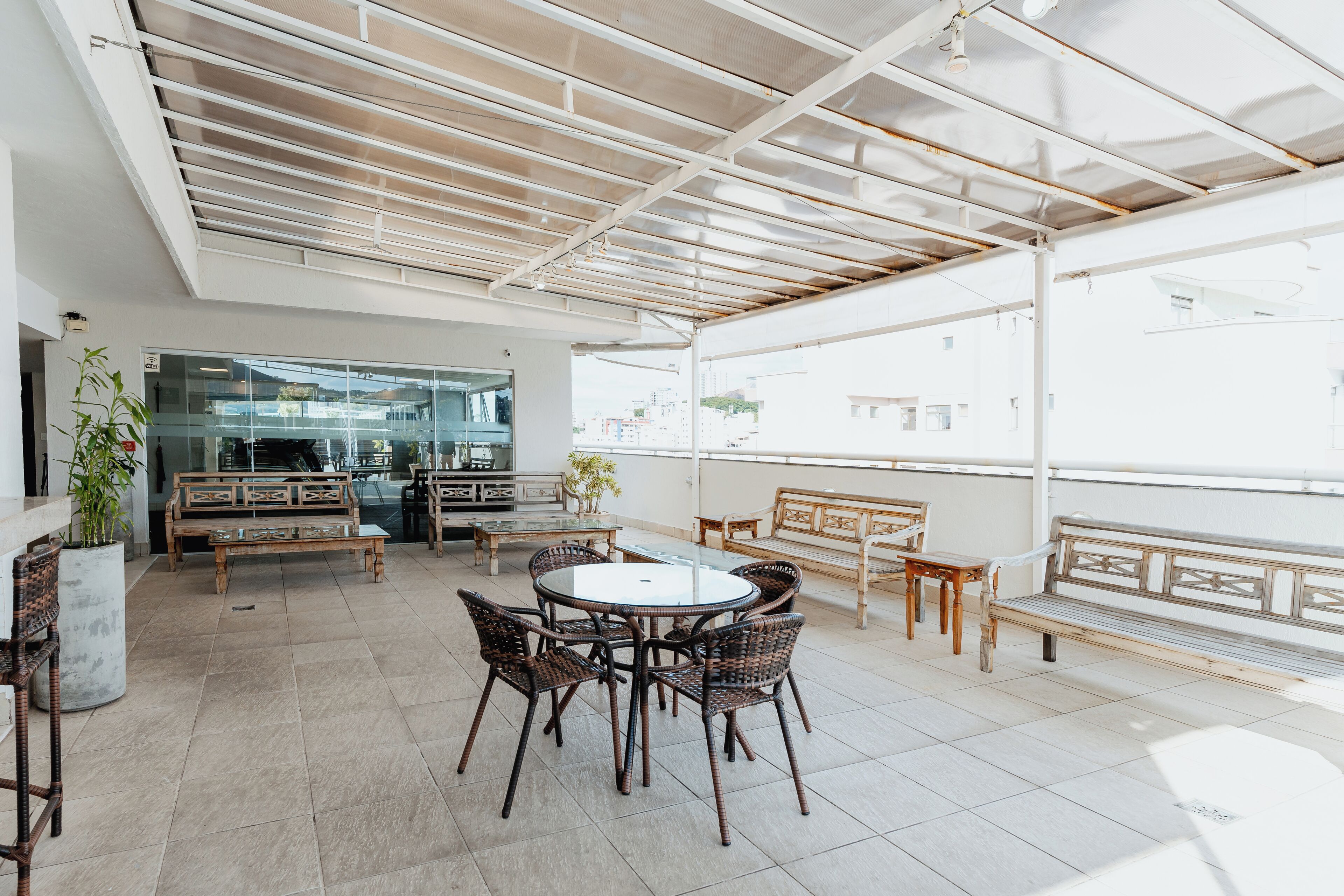 A covered outdoor seating area with a round table and wicker chairs, a glass wall with a view of buildings, and a wooden bench along the railing.