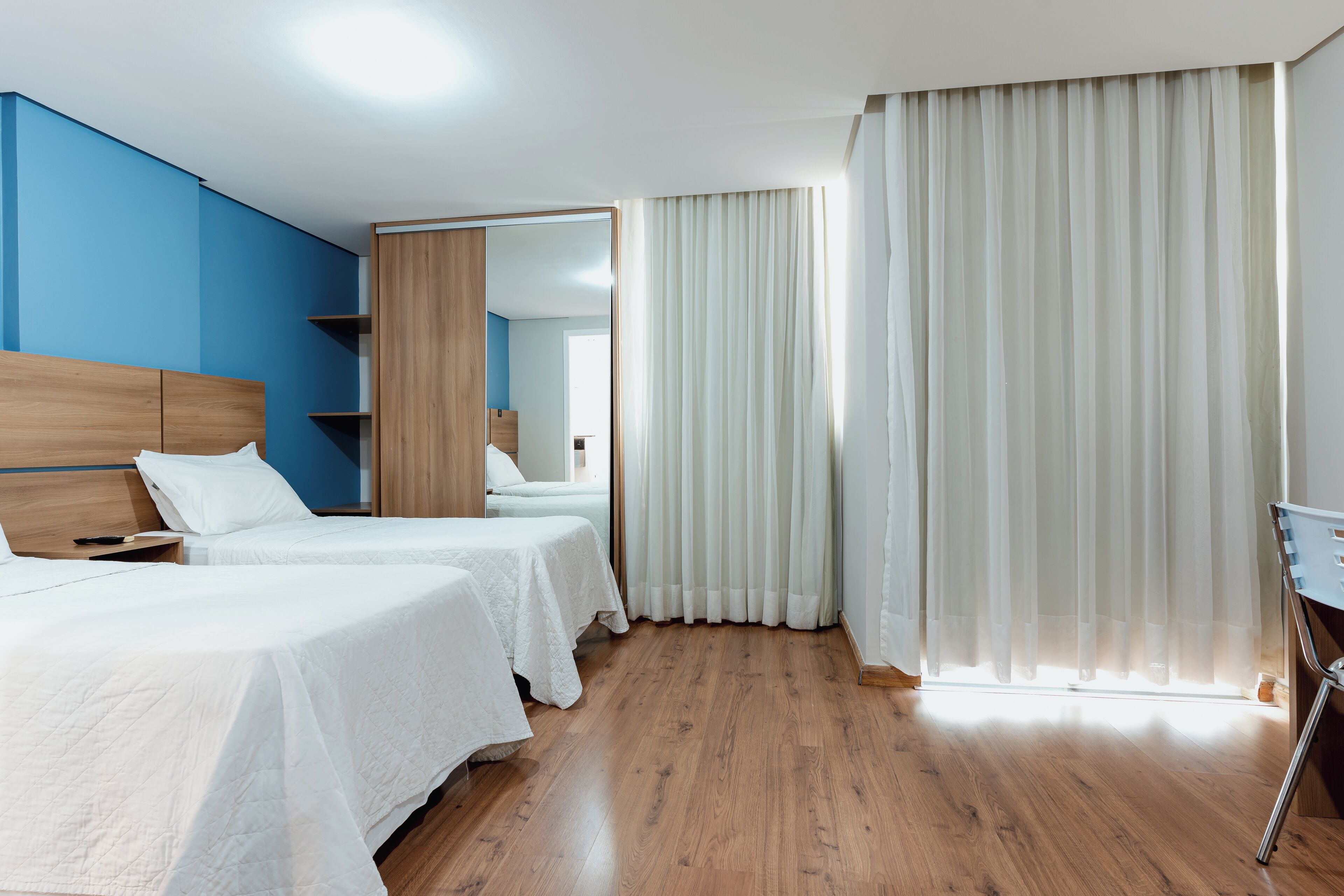 Habitación de hotel con dos camas, cabeceros de madera, un espejo grande y vista a la calle a través de cortinas translúcidas.