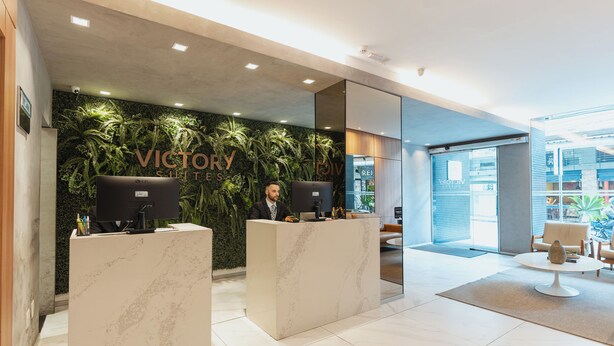 A reception area with a marble counter, a receptionist, and a wall decorated with green plants. - Victory Suites (Juiz de Fora)