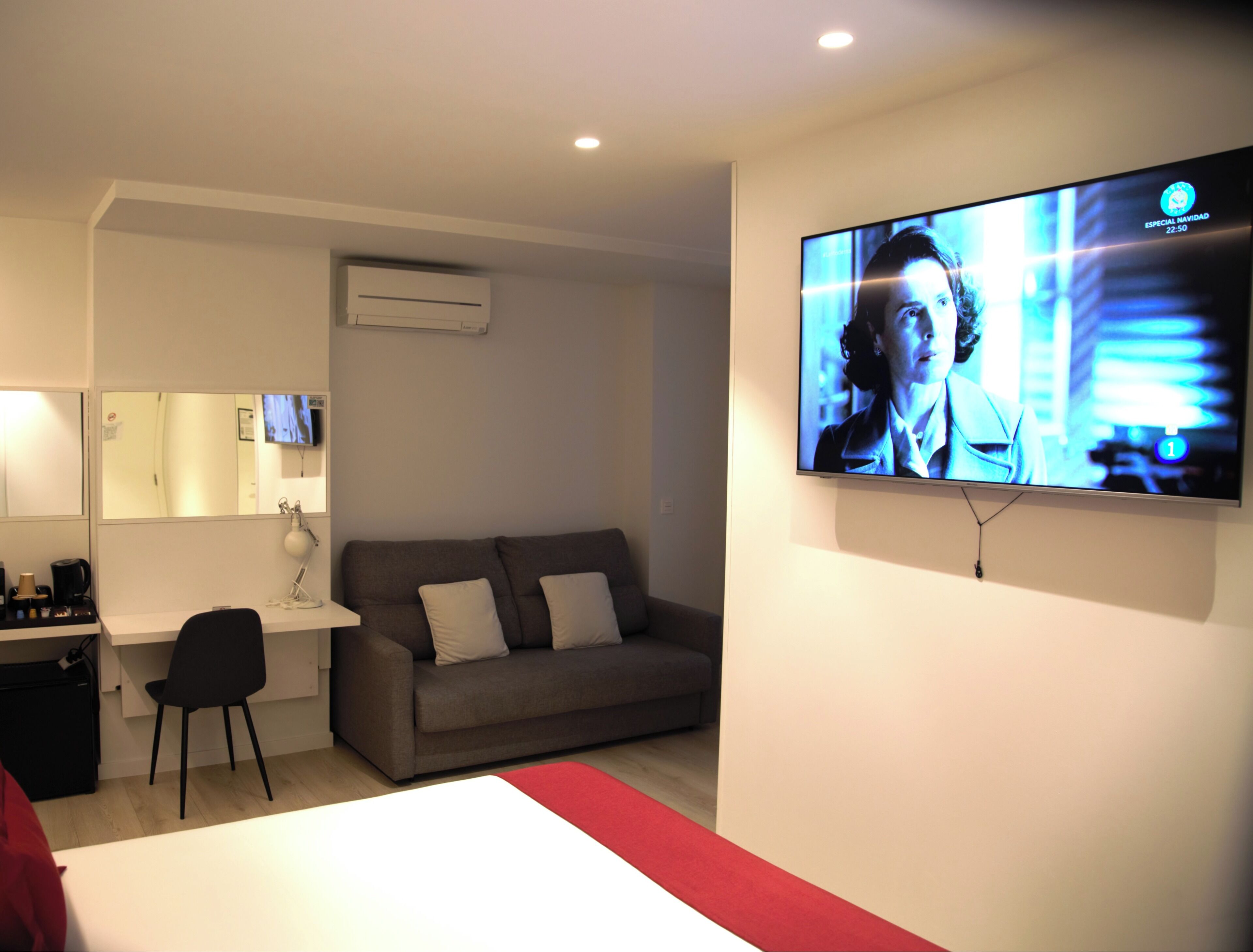 A modern hotel room with a flat-screen TV mounted on the wall, a grey sofa, a desk with a chair, and a bed with a red accent.