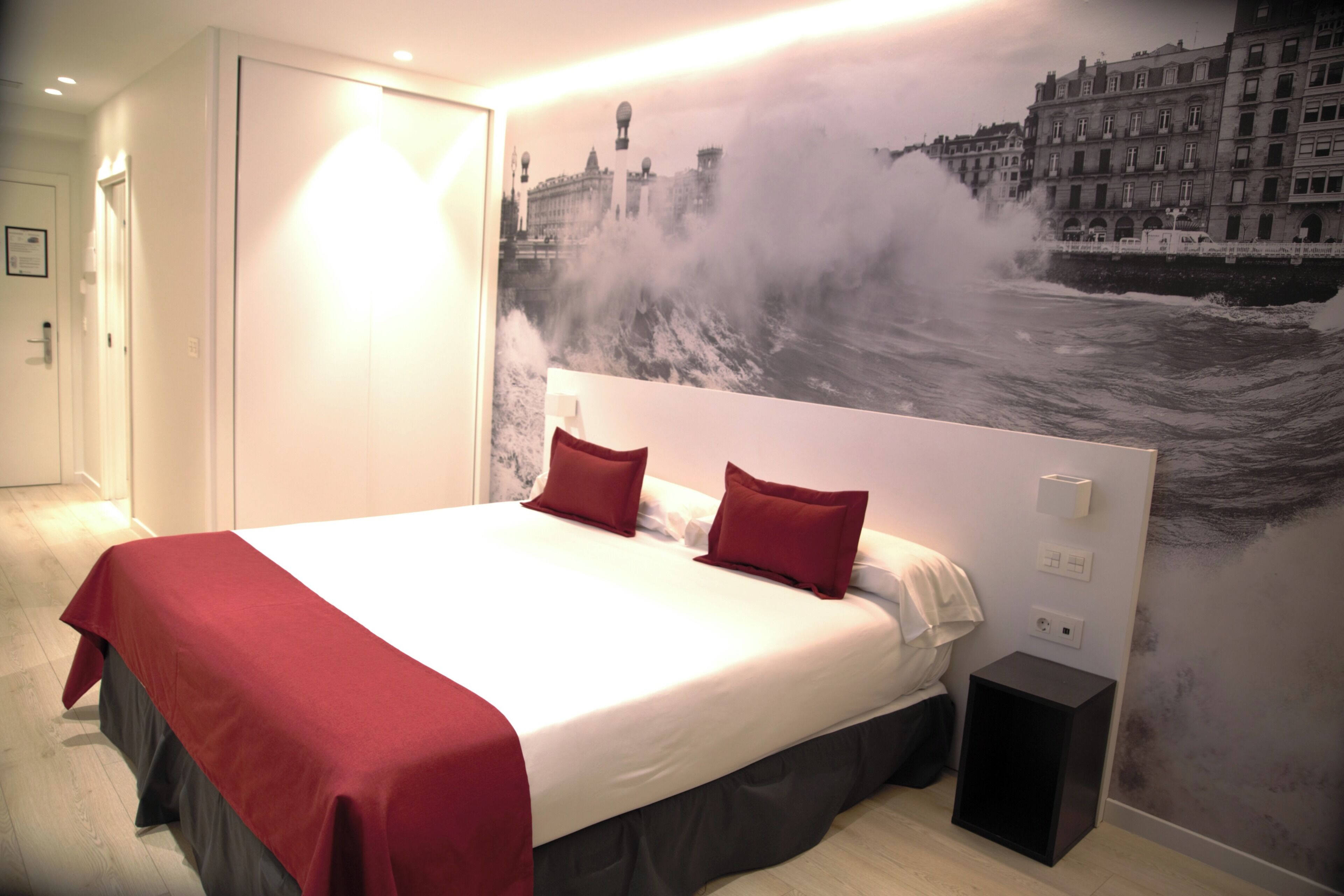 A hotel room with a large bed, red bedding, a wall mural of a cityscape, and a view of the sea.