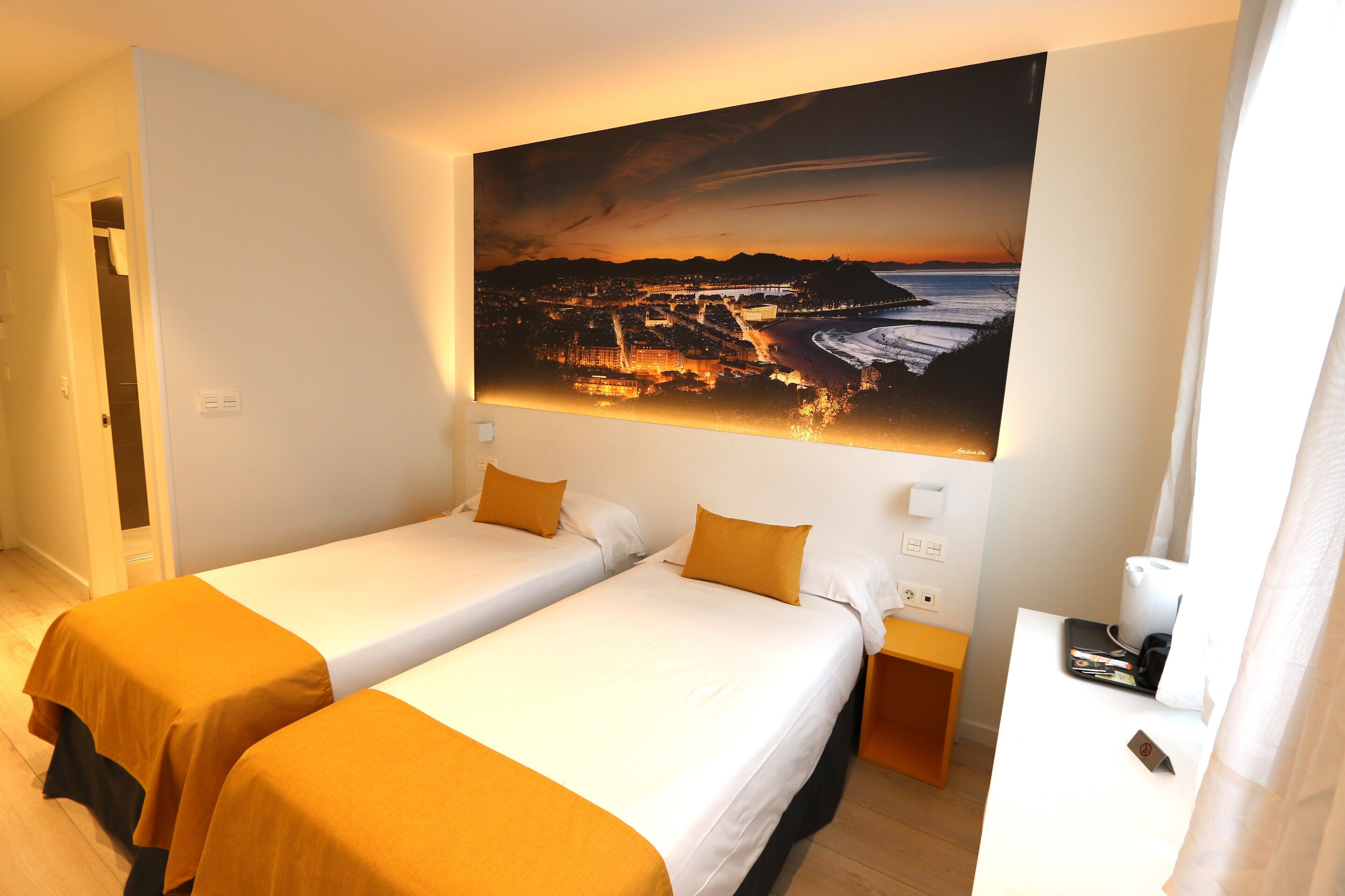 Een hotelkamer met twee bedden, een schilderij van een kuststadsgezicht, een bureau met telefoon en koffiezetapparaat, en een deur die leidt naar een kast.