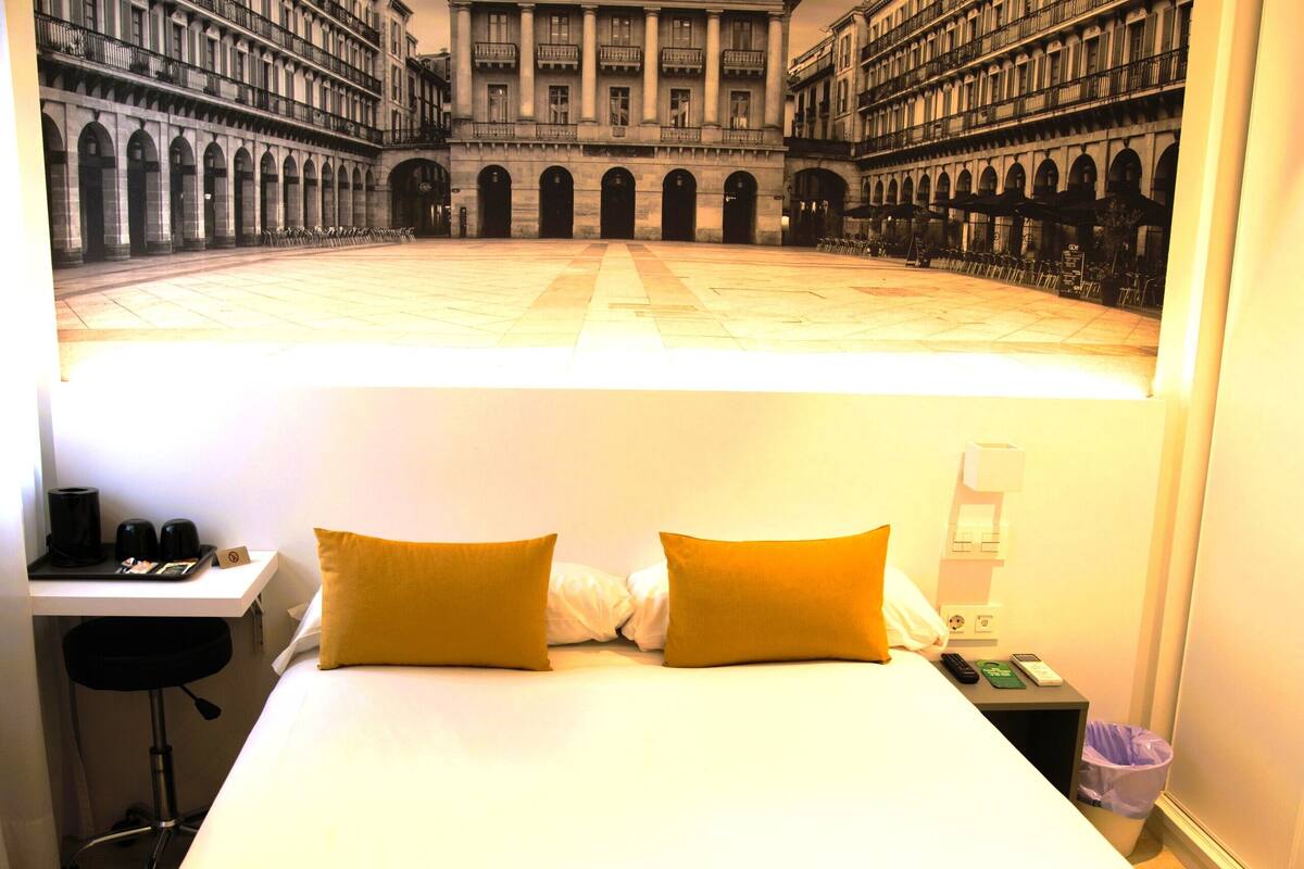 Quarto de hotel com uma cama, uma escrivaninha e uma mesa de cabeceira.