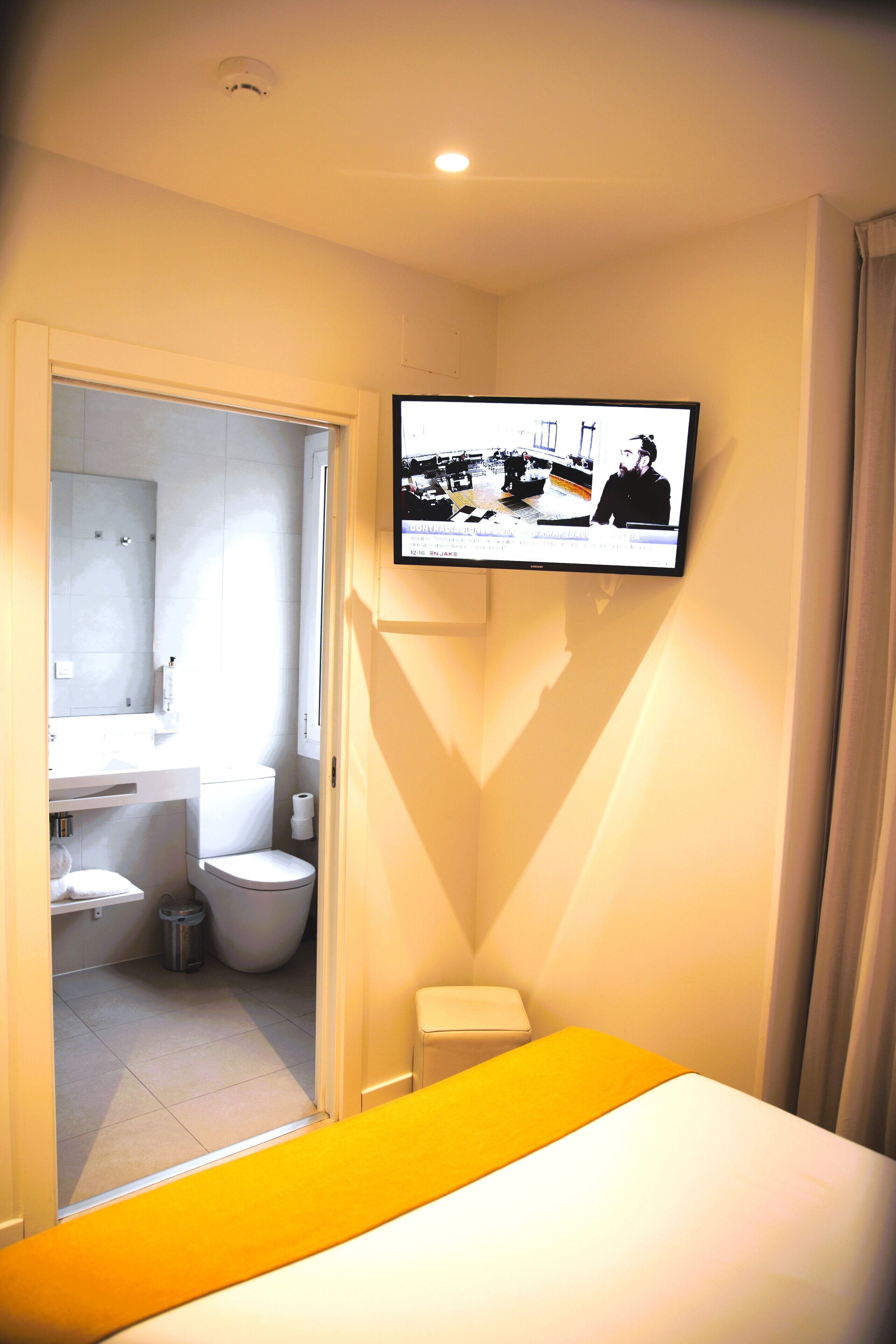 Een hotelkamer met een bed, een aan de muur bevestigde televisie, een badkamer met toilet en wastafel, en een kleine prullenbak.